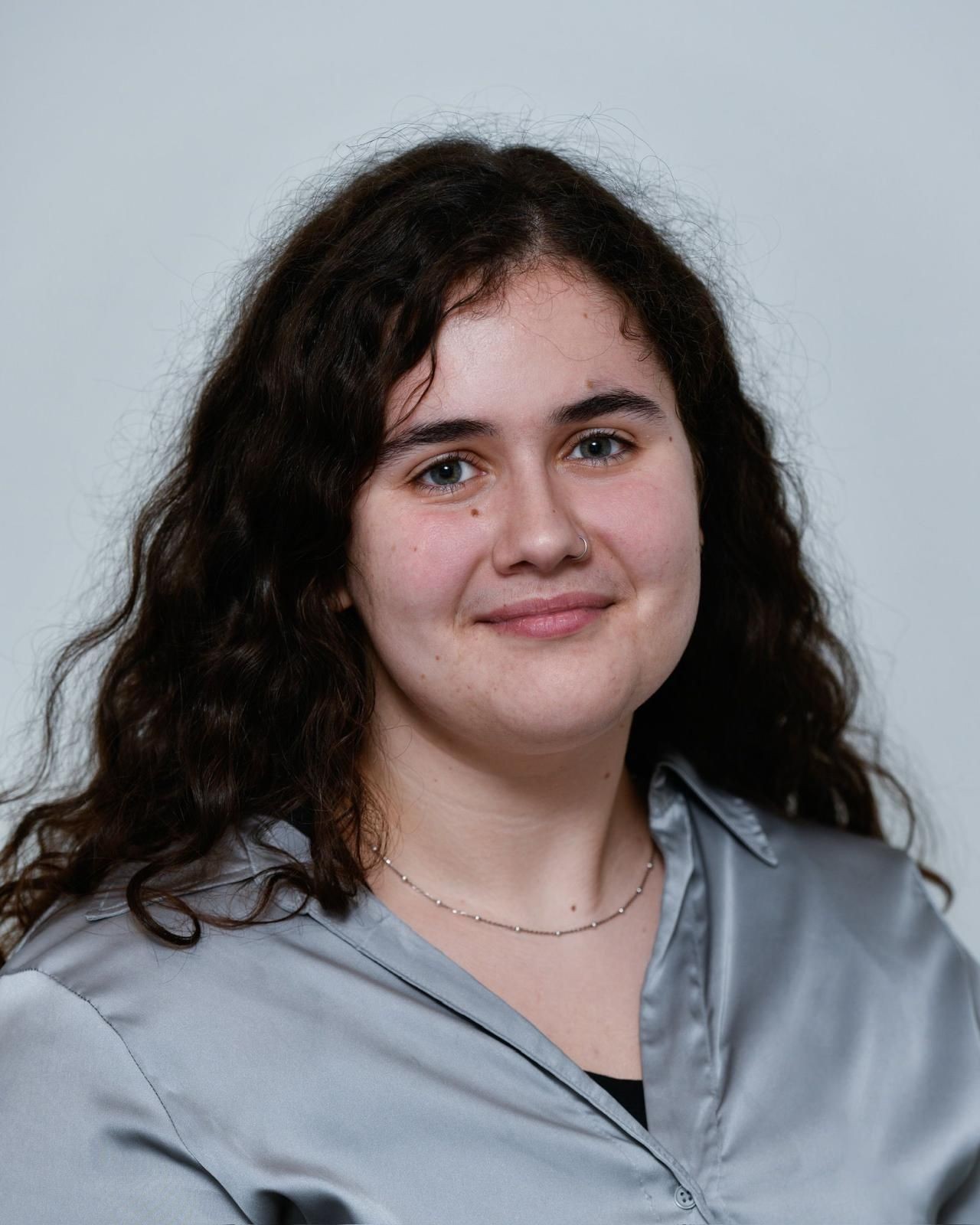 Person with long curly hair wearing a gray silk shirt and a delicate necklace.