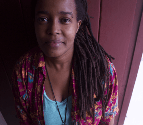 Person wearing a colorful floral shirt and necklace with long dreadlocks standing against a maroon wall.